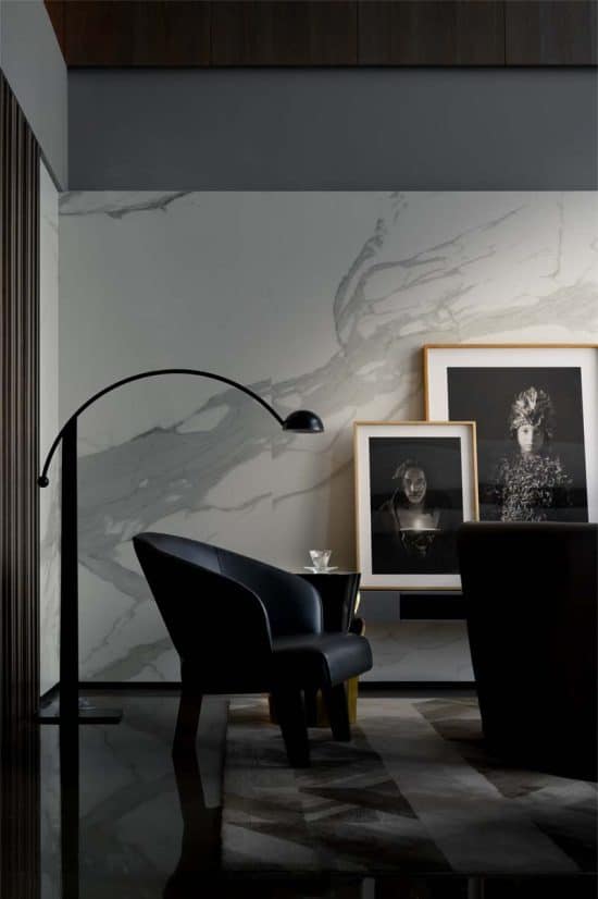 A modern, minimalist living space reminiscent of chic hotels in London features a sleek, black floor lamp arching over a dark armchair. A small side table with a glass sits beside the chair. The marble-textured wall behind displays framed monochrome portraits, adding an artistic touch to the room.