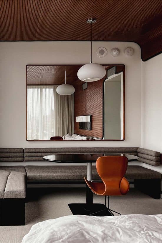 A contemporary hotel room in London with modern furniture. The room features a brown wood-paneled ceiling, a large rectangular mirror, a round black table with an orange chair, and a built-in gray bench with cushions. A bed with white linens is visible in the foreground.