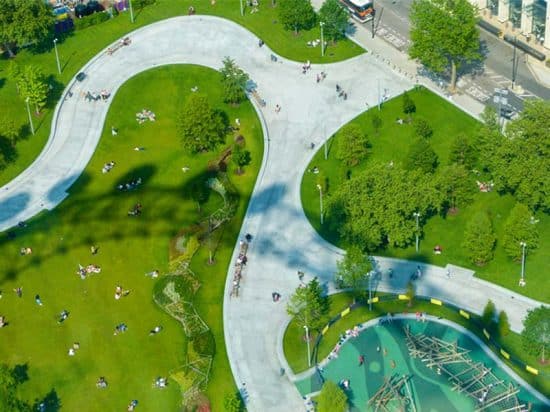 Aerial view of a park with winding paths and green lawns. People are scattered around, sitting and walking. The park features several trees, a pond with wooden structures, and nearby roads with light traffic and buildings, reminiscent of the serene surroundings near hotels in London. The scene is sunny and vibrant.