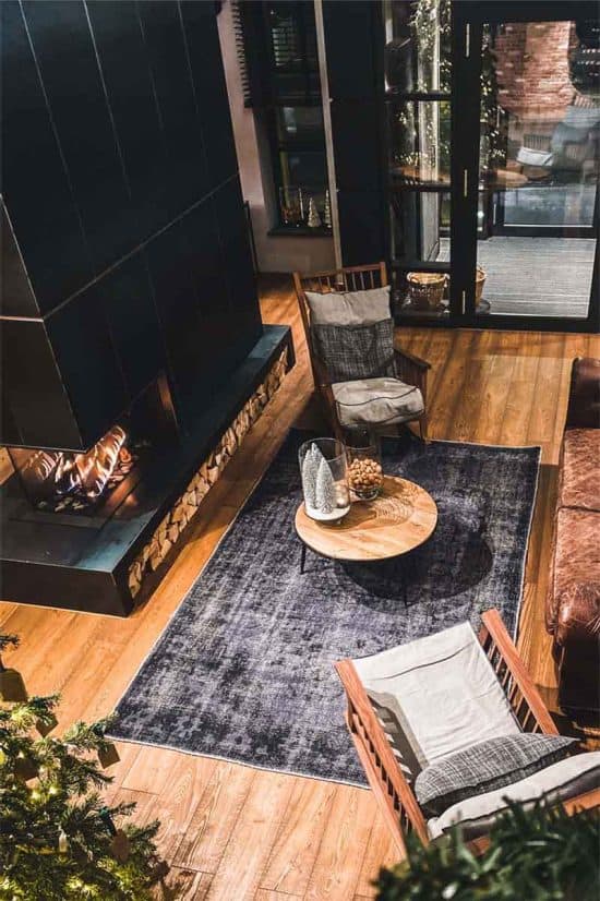 A cozy modern living room with a lit fireplace built into a black wall evokes the comfort of boutique hotels in London. Two wooden armchairs with cushions and a small wooden round table with a decorative centerpiece are placed on a dark gray rug. Glass doors open to a patio area visible outside.