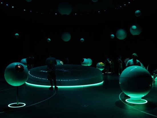 A dimly lit, futuristic exhibit in one of the renowned hotels in London features glowing, spherical displays and a large illuminated circular table at its core. People are examining the interactive installations, which emit a soft green light and create an otherworldly atmosphere.