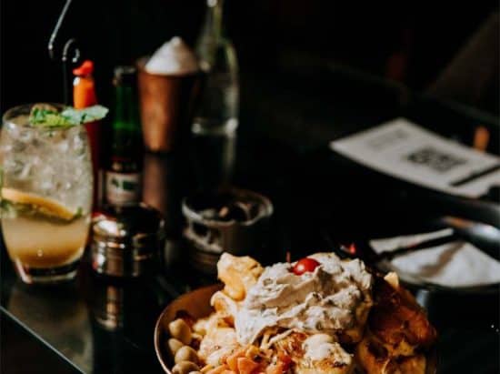 A plate of nachos topped with sour cream, cheese, and tomatoes sits on a glass table. Next to it, there's a refreshing beverage garnished with mint and a slice of lemon. Condiments, a pepper mill, and a menu are visible in the background—an inviting scene reminiscent of fine dining at hotels in London.