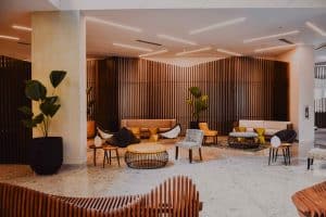 A modern lobby area with stylish furniture, including sofas, chairs, and round tables. The space features wooden slat walls, large potted plants, and recessed lighting. The floor is tiled, and the decor is a mix of neutral and vibrant colors typical of upscale hotels in London.