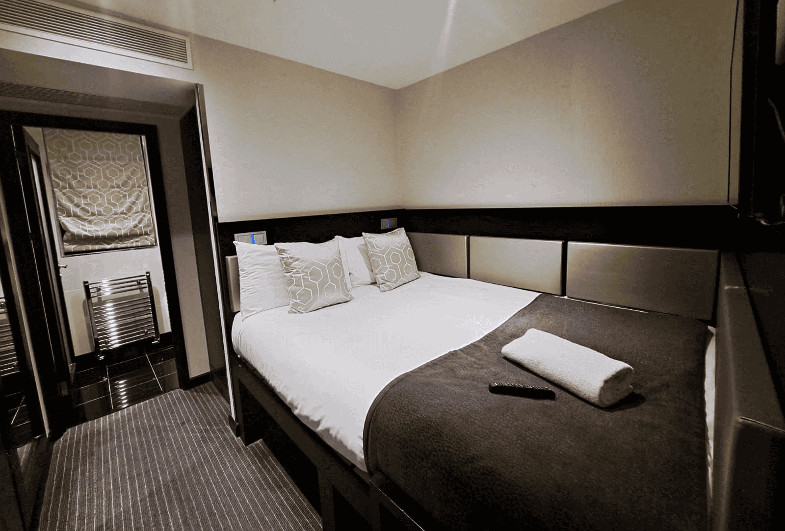 A cozy, modern hotel room featuring a neatly made double bed with white linens and decorative pillows. A folded towel and a TV remote rest on the bed. A window with a patterned blind and a small bathroom with a towel rack can be seen in the background.