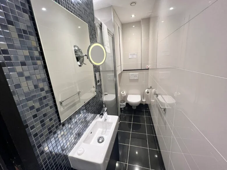 A modern bathroom featuring white tiled walls and a black tiled floor. The room includes a small white sink with a mirror above it, a wall-mounted toilet, and a shower area with glass partition. The walls have reflective mosaic tiles.