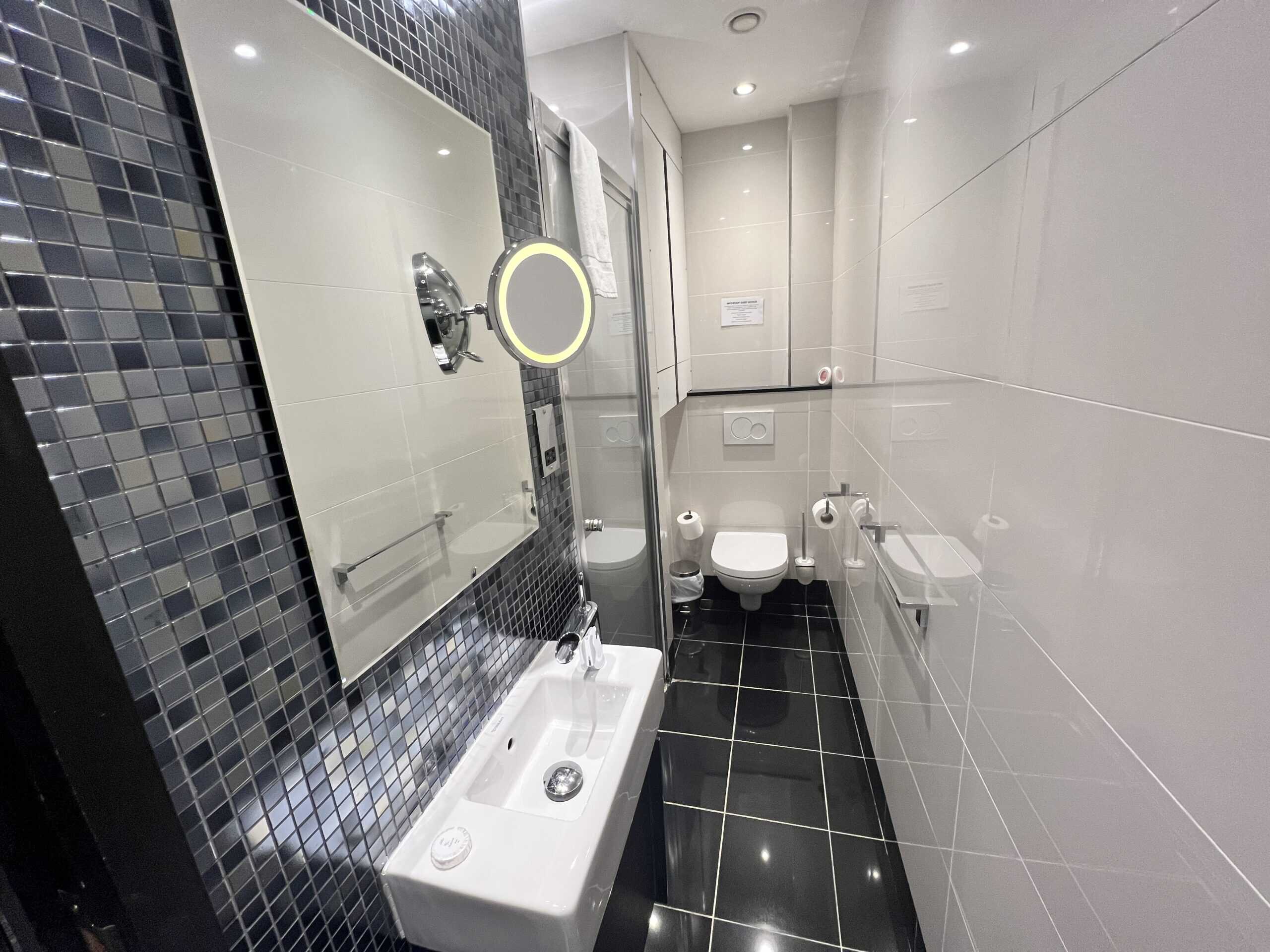 A modern bathroom featuring white tiled walls and a black tiled floor. The room includes a small white sink with a mirror above it, a wall-mounted toilet, and a shower area with glass partition. The walls have reflective mosaic tiles.