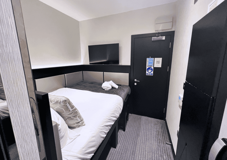 A compact hotel room features a neatly made bed with white linens and decorative pillows against a wall. A mounted flat-screen TV is positioned above the bed's headboard. The room also includes a black door with various signs and a small closet-like area.