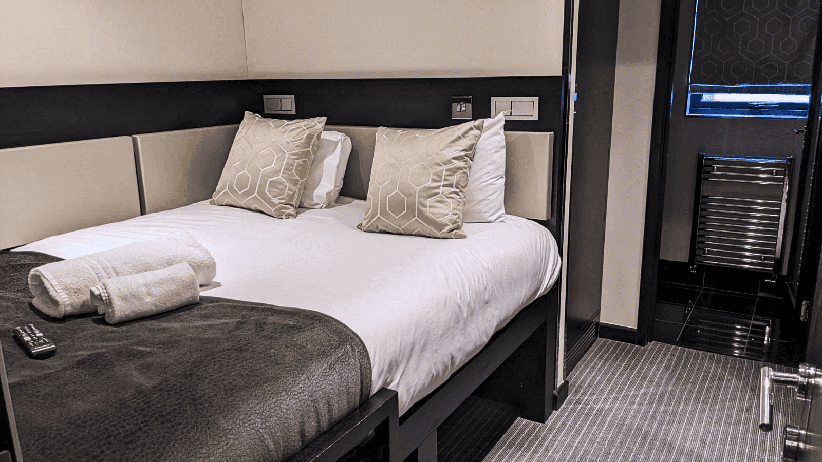 A modern hotel room with a neatly made double bed featuring beige and white pillows, and a folded white towel on top. A remote control lies on a dark coverlet. The room has neutral colors with a window in the back and a bathroom visible through a door.