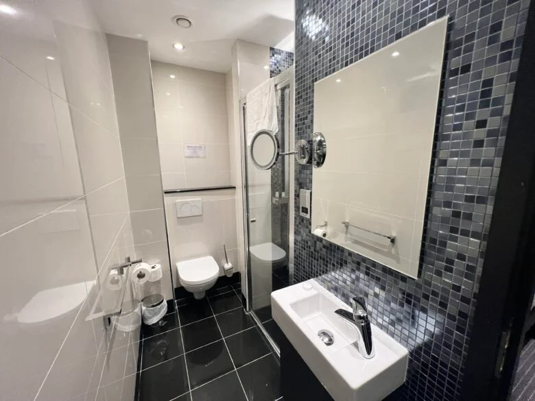 A modern bathroom with a white toilet, a wall-mounted sink, and a walk-in shower. The walls are tiled with white and black mosaic tiles, and the floor has large black tiles. A large mirror, a circular magnifying mirror, and various bathroom fixtures are present.