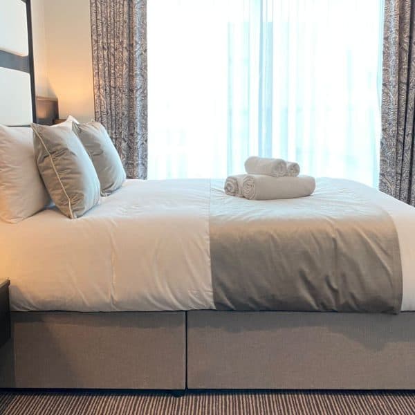 A neatly made double bed in a modern hotel room with neutral decor at one of the finest hotels in London. The bed has several pillows and two sets of rolled towels on top. A nightstand with a lamp and telephone stands beside the bed, and a large window with curtains lets in natural light.