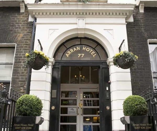 The entrance of Arran Hotel, one of the charming hotels in London, is marked by a tall arch labeled "ARRAN HOTEL 77." Flanking the entrance are two hanging flower baskets and two potted shrubs, with black railings on either side. The facade is a mix of white and dark brick.