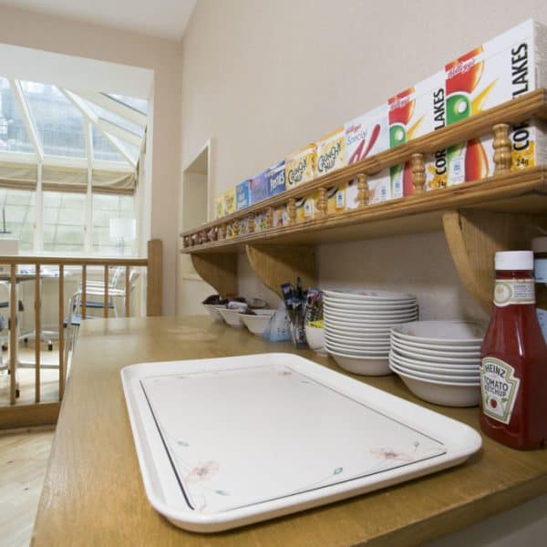 A kitchen area featuring a wooden counter with a white serving tray, a variety of cereals displayed on a shelf above, and condiments including ketchup and HP sauce on the side. In the background, there's a dining area with tables and chairs near large windows, evoking the charm of hotels in London.