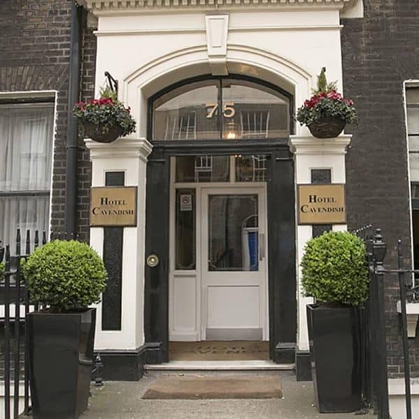 Entrance of Hotel Cavendish, one of the distinguished hotels in London, featuring a white door with the number 75 above it, flanked by two signs reading "Hotel Cavendish." Two large potted plants with manicured greenery stand on either side, and hanging flower baskets adorn the facade.