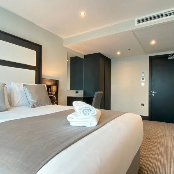 A modern hotel room in one of the finest hotels in London, featuring a neatly made double bed with gray and white linens and two folded towels on top. The room includes a desk with a chair, a flat-screen TV mounted on the wall, and a black door in the background.