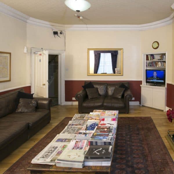 A cozy living room with beige walls and a wooden floor replicates the charm often found in hotels in London. It features a large brown leather sofa and loveseat. A coffee table in the center holds various magazines. A TV in the corner is on, and a fireplace with a mirror above it is on the right side.