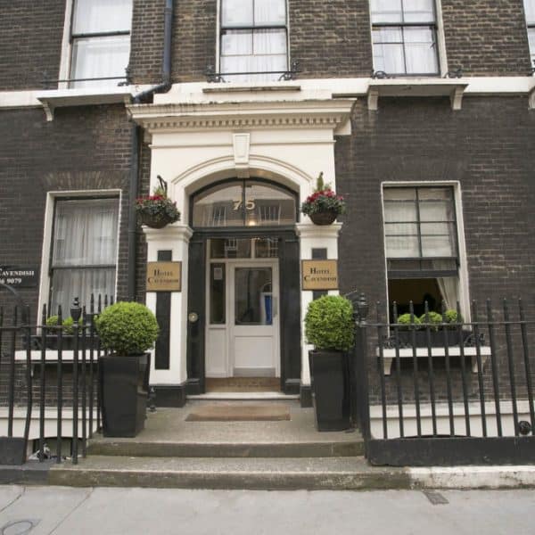 Front view of a brick building with a white door surrounded by a grand white frame. Potted plants line the entrance. Two signs next to the door read "Hotel Cavendish," making it one of the charming hotels in London. The building features multiple windows with white frames and black railings in front.