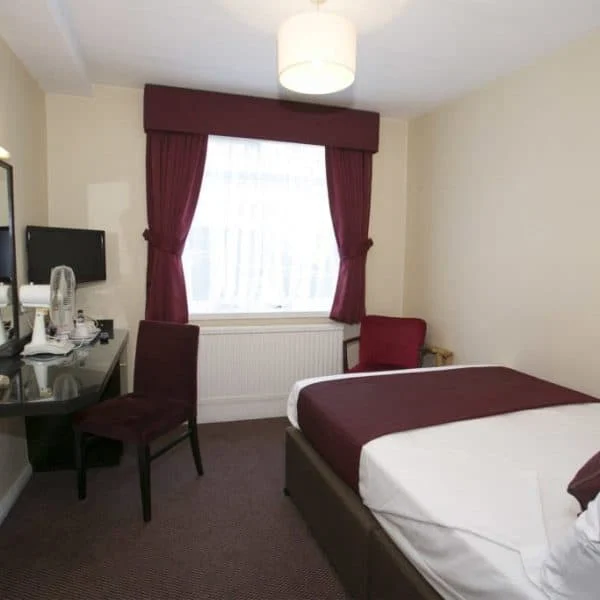 A neatly arranged hotel room in London features a single bed with maroon bedding and pillows. A matching maroon curtain covers the window. A work desk with a chair, a wall-mounted telephone, and a small television adorn the room. A maroon armchair sits in the corner.