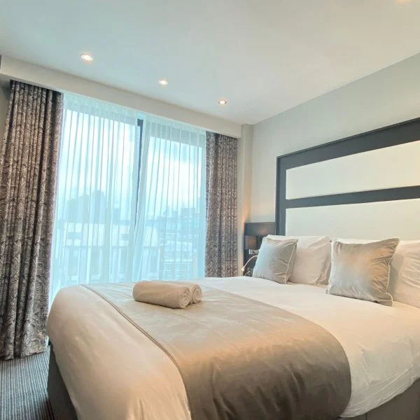 A modern hotel room in one of the finest hotels in London features a large bed with gray and white bedding, three pillows, and a folded towel. Two bedside tables with lamps flank the bed. A large window with sheer and patterned curtains allows natural light to fill the space.