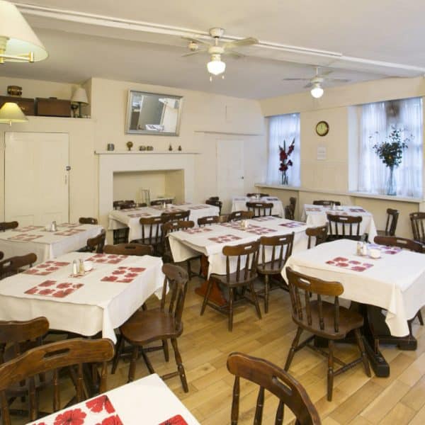 A cozy indoor dining room at one of the finest hotels in London, featuring wooden chairs and tables covered with white tablecloths and red placemats. The room has hardwood flooring and light-colored walls, complemented by wall-mounted light fixtures and a small plant hanging by the window.