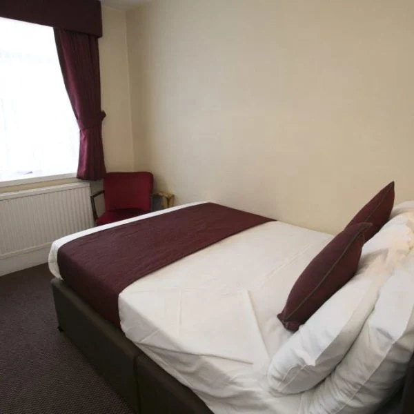 A small, cozy bedroom with a double bed covered in white linens and a maroon blanket and pillows. The room has beige walls, a large window with maroon curtains, and a red armchair in the corner, with a radiator under the window. The floor is carpeted—reminiscent of charming hotels in London.