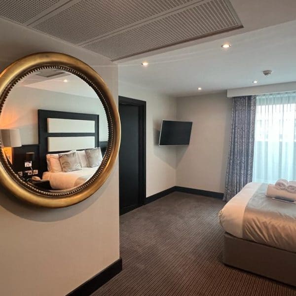 A modern hotel room in one of the best hotels in London features a large, circular mirror on the wall, reflecting a neatly made bed with pillows. The room includes a wall-mounted TV, a large window with sheer curtains, and a neatly folded towel on the bed. The decor is minimalist with neutral tones.