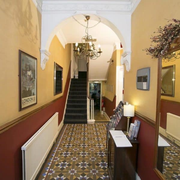 The image shows a hallway with patterned tile flooring and red and yellow walls, reminiscent of the charming decor often found in hotels in London. A staircase at the end leads up, while a chandelier hangs from a white decorative ceiling. There's a mirror, framed artwork on the walls, and a wooden side table holding brochures.