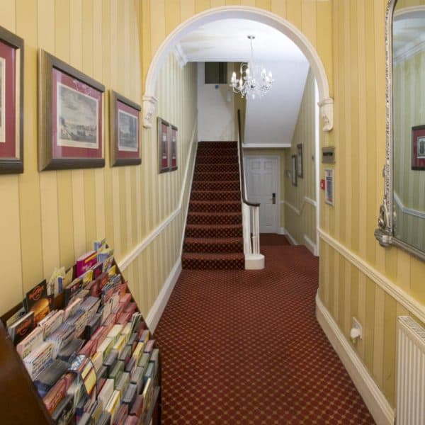 A narrow hallway with striped yellow walls leads to a staircase covered in red carpet. A chandelier hangs from the ceiling, reminiscent of classic hotels in London. There's a large mirror on the right wall and a rack of brochures on the left side. The staircase leads to an upper floor and a white door.