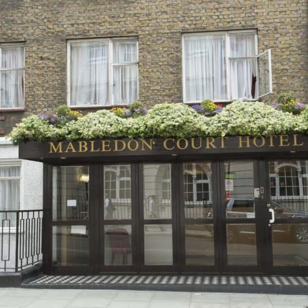 The image depicts the entrance of the Mabledon Court Hotel, one of the prominent hotels in London. The facade features large glass doors and windows with a black awning displaying the hotel's name in gold letters. Above the awning are flower boxes with blooming plants, while a sidewalk runs in front of the entrance.