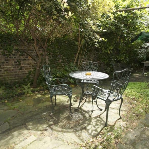 A serene garden scene features a round metal table with four matching chairs. The area is shaded by leafy trees and surrounded by a brick wall covered in ivy. Sunlight filters through the foliage, creating a peaceful and inviting atmosphere reminiscent of hidden gems in hotels in London.