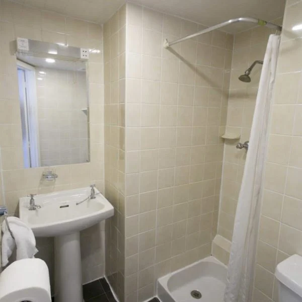 A small bathroom with beige tiled walls and a black tiled floor, reminiscent of chic hotels in London. It features a white pedestal sink with a wall-mounted mirror, a white toilet with a roll of toilet paper attached to the wall, and a corner shower with a white curtain.