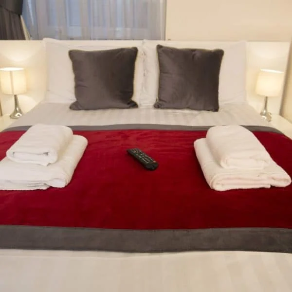 A neatly made bed with white sheets and a red throw blanket, topped with two folded white towels and a TV remote control placed in the center. The bed is flanked by two lit bedside lamps. There are two gray pillows propped up against the headboard, offering a welcoming touch reminiscent of hotels in London.