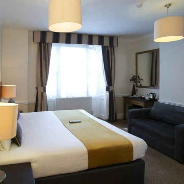 A modern hotel room in one of the chic hotels in London, featuring a king-sized bed with two bedside lamps and a striped headboard adorned with a mustard-colored runner. A dark sofa sits against one wall, facing a dresser with a mirror, plus a flat-screen TV and tea/coffee station are visible.