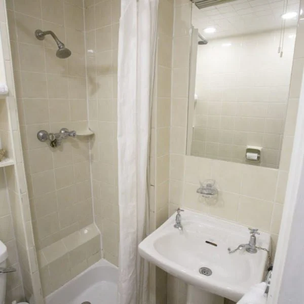 A compact bathroom with beige tiles, reminiscent of those found in quaint hotels in London, features a small shower stall with a curtain, a white pedestal sink with two faucets, a mirror above the sink, and a toilet partially visible on the left. A towel hangs on a rack beside the sink.