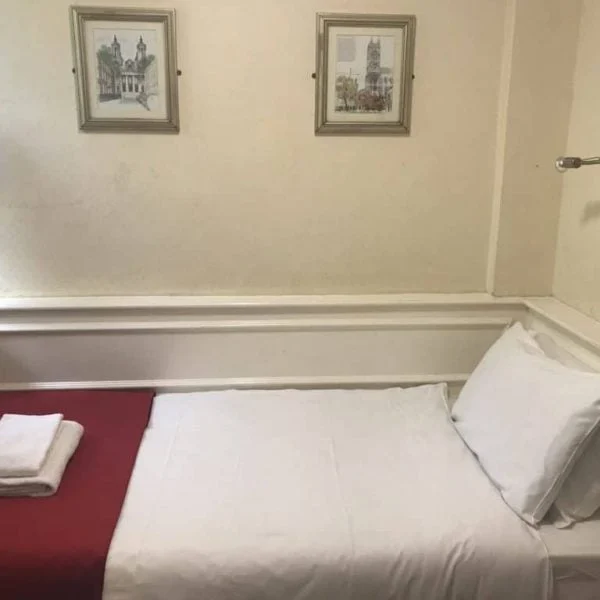 A small, tidy hotel room with a single bed in the center. The bed is made with white linens and has a red throw at the foot, topped with neatly folded towels. The walls are decorated with three framed pictures, and a wall-mounted reading light is positioned above the bed—one of the charming hotels in London.