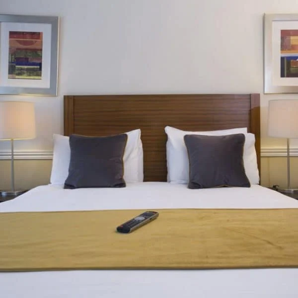 A neatly made bed with a wooden headboard, two black pillows, and a yellow runner across the foot of the bed evokes the cozy ambiance of upscale hotels in London. Two bedside tables each hold a lamp, and two framed artworks hang above the headboard. A TV remote is placed on the yellow runner.