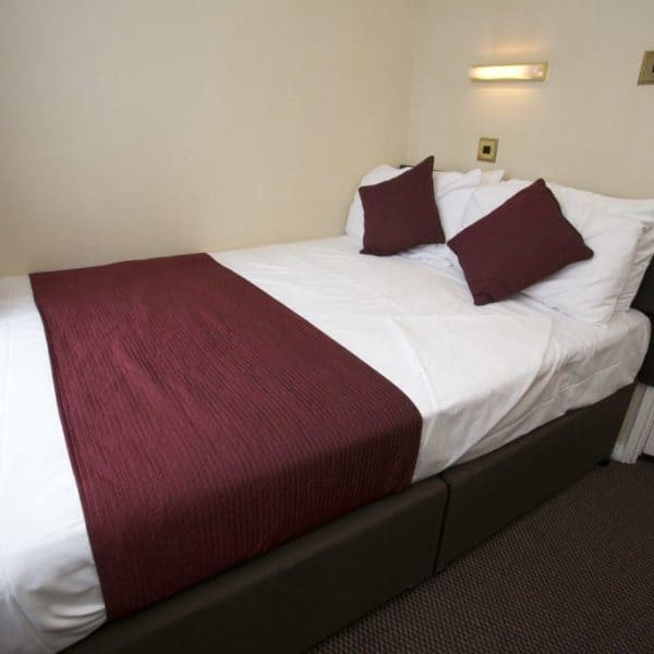 A small neatly made bed with white linens, a dark purple throw, and two matching pillows is situated against a light-colored wall. A wall-mounted reading light is above the bed, and a window with a purple curtain is on the left side. This cozy setup evokes the charm of fine hotels in London.