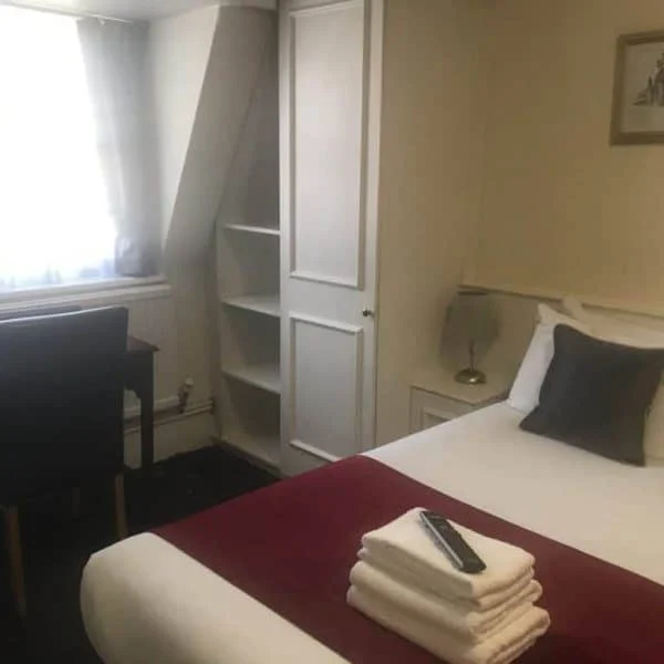 A cozy bedroom in one of the finest hotels in London features a neatly made bed with white sheets, a burgundy throw, and stacked towels with a TV remote on top. The room includes a desk and chair near the window with curtains partially drawn. A small built-in wardrobe is visible next to a wall-mounted picture.