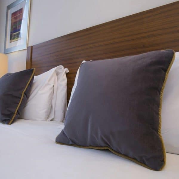 A neatly made bed with white linens, two large dark gray pillows with gold trim, and a polished wooden headboard. A softly lit lamp and framed painting are in the background, partially visible on the left side, emulating the elegance often found in hotels in London.