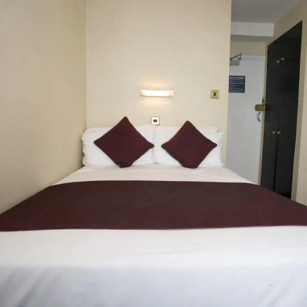 A small, neatly arranged hotel room with a double bed centered against a beige wall. The bed has white linens, a burgundy throw blanket, and two matching burgundy pillows. A wall-mounted lamp is above the bed, and a door and dark-colored wardrobe are visible in the background—perfect for those seeking hotels in London.