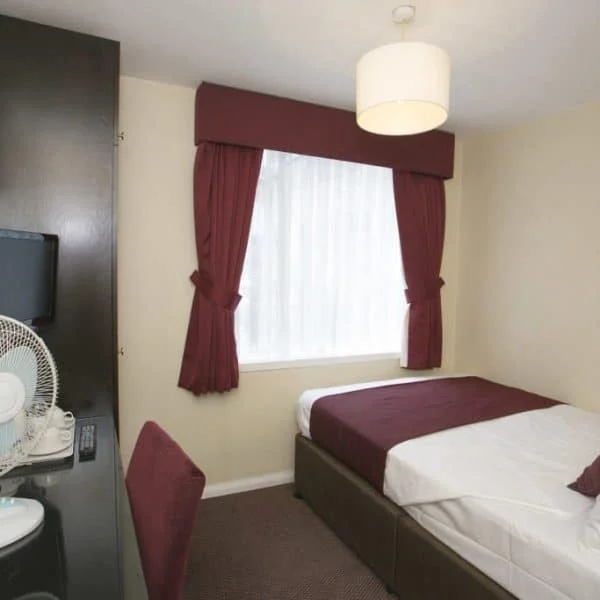 A small, cozy hotel room in London features a bed with white and burgundy bedding, a window with burgundy drapes, a wall-mounted flat screen TV, a dresser with a mirror and a fan, and a brown desk chair. The room has neutral-colored walls and brown carpet.