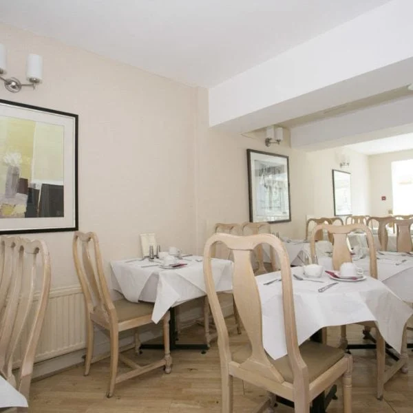 A small, well-lit dining area with several wooden tables and chairs. Each table is neatly set with white tablecloths and tableware. Abstract art paintings adorn the light-colored walls, and natural light streams in through large windows, reminiscent of the cozy dining spaces found in hotels in London.