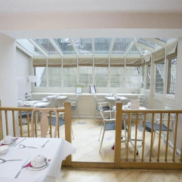 A cozy dining area featuring a small number of tables set with cups and saucers, located in a sunroom with large windows and a glass ceiling. A wooden railing separates this space from the main room. Light wooden flooring and neutral-colored walls create a bright atmosphere, reminiscent of quaint hotels in London.