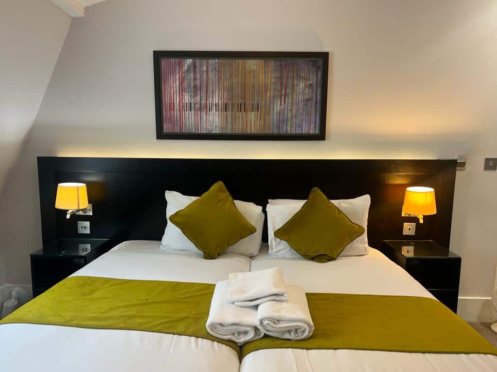 A neatly made bed in a modern hotel room with a black headboard, two green cushions, and a green bed runner. Two folded towels are placed on the bed. Bedside tables with orange lamps flank the bed, and a colorful abstract painting hangs above the headboard.