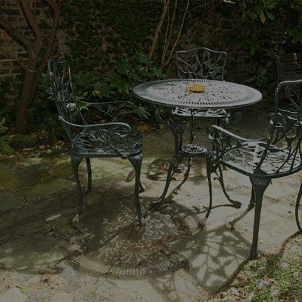 An outdoor garden scene at one of the finest hotels in London features a round, ornate metal table and four matching chairs on a stone-paved area. The garden is lush with greenery and vines climbing a brick wall in the background. Sunlight casts intricate shadows from the furniture onto the ground.