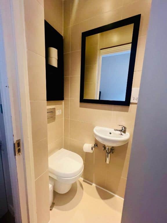 A small, beige-tiled bathroom featuring a wall-mounted toilet with a toilet paper holder to the right. Above the toilet is a shelf with toilet paper rolls. Opposite the toilet is a small white wall-mounted sink with a mirror above it.