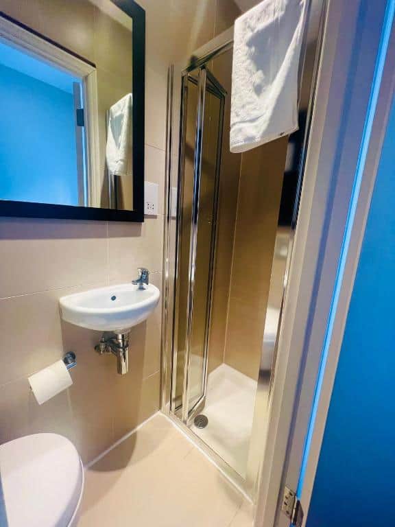 A compact bathroom featuring a small sink with a wall-mounted mirror above it, a modern shower with a glass door, and a white towel hanging on the shower frame. There is a toilet partially visible with a toilet paper holder attached to the wall beside it.