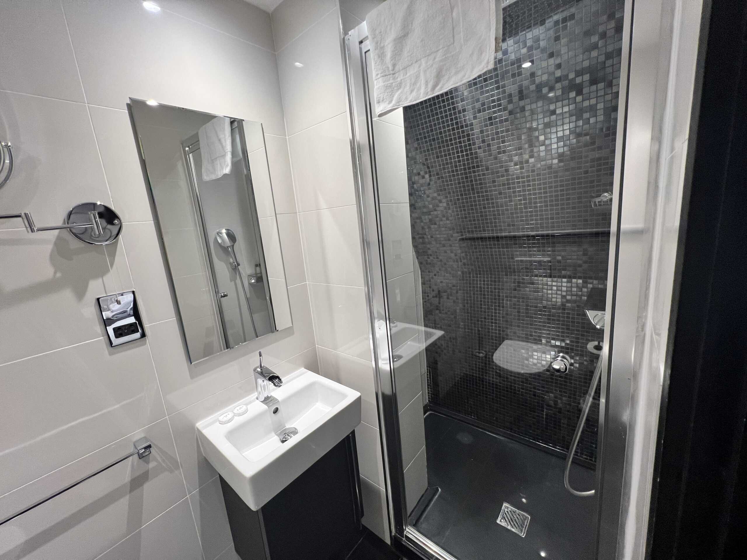 A modern bathroom with grey tiled walls, featuring a sink with a square mirror above it on the left side, and a walk-in shower with a glass door and dark mosaic tiles on the right. A towel hangs over the shower door.