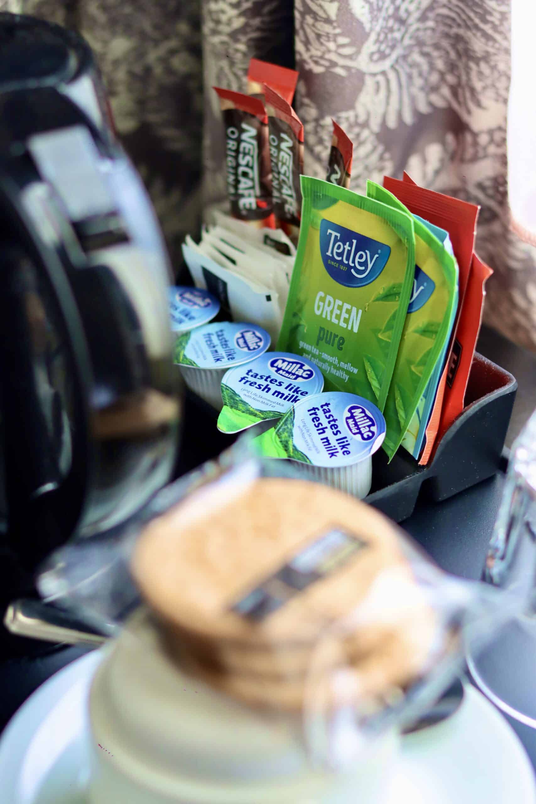 A close-up view of a hotel room's coffee and tea station. The tray includes Nescafé instant coffee packets, Tetley green tea bags, single-serve creamers, a stack of disposable cups with lids, and a coffee machine, set against a background with patterned curtains.