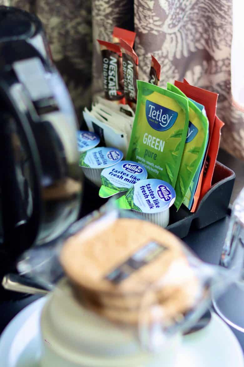 A close-up view of a coffee and tea station featuring Tetley green tea bags, Nescafé instant coffee sachets, creamers, and sugar packets, all neatly arranged on a tray. A coffee maker and a cup with a wooden lid are visible in the foreground.