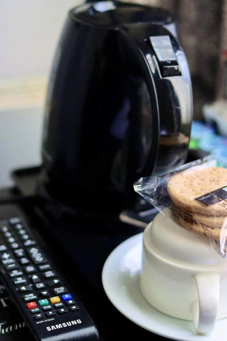 A close-up of a black electric kettle on a counter. In front of the kettle is a white cup and saucer with a clear package of biscuits resting on top. A TV remote control lies next to the cup on the left side.